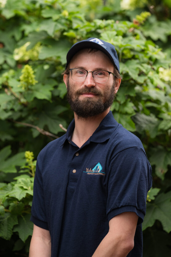 Portrait of a bearded man with glasses wearing a navy polo shirt and cap, posing outdoors among lush green foliage.
