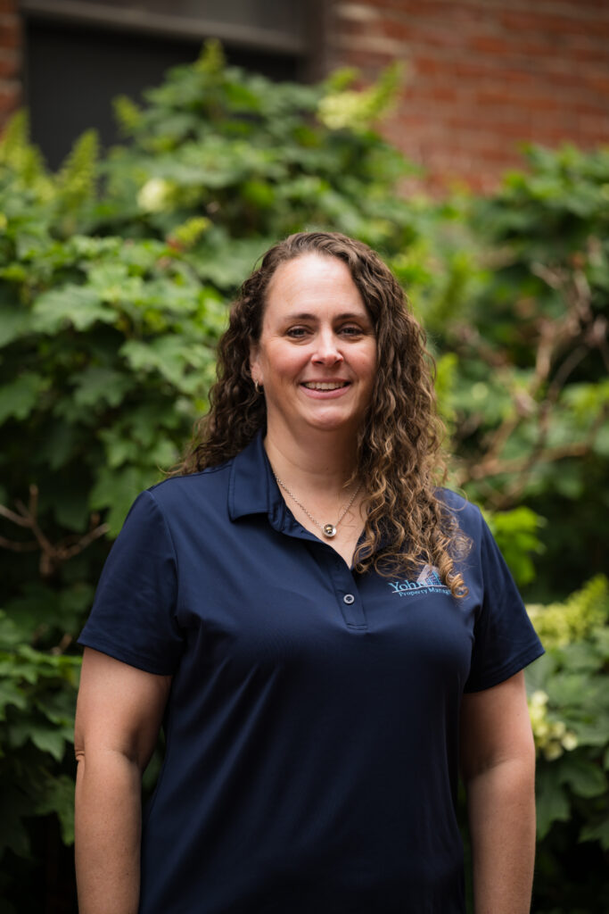Smiling woman with curly brown hair standing outdoors in front of greenery, wearing a navy polo shirt with a logo on the chest.